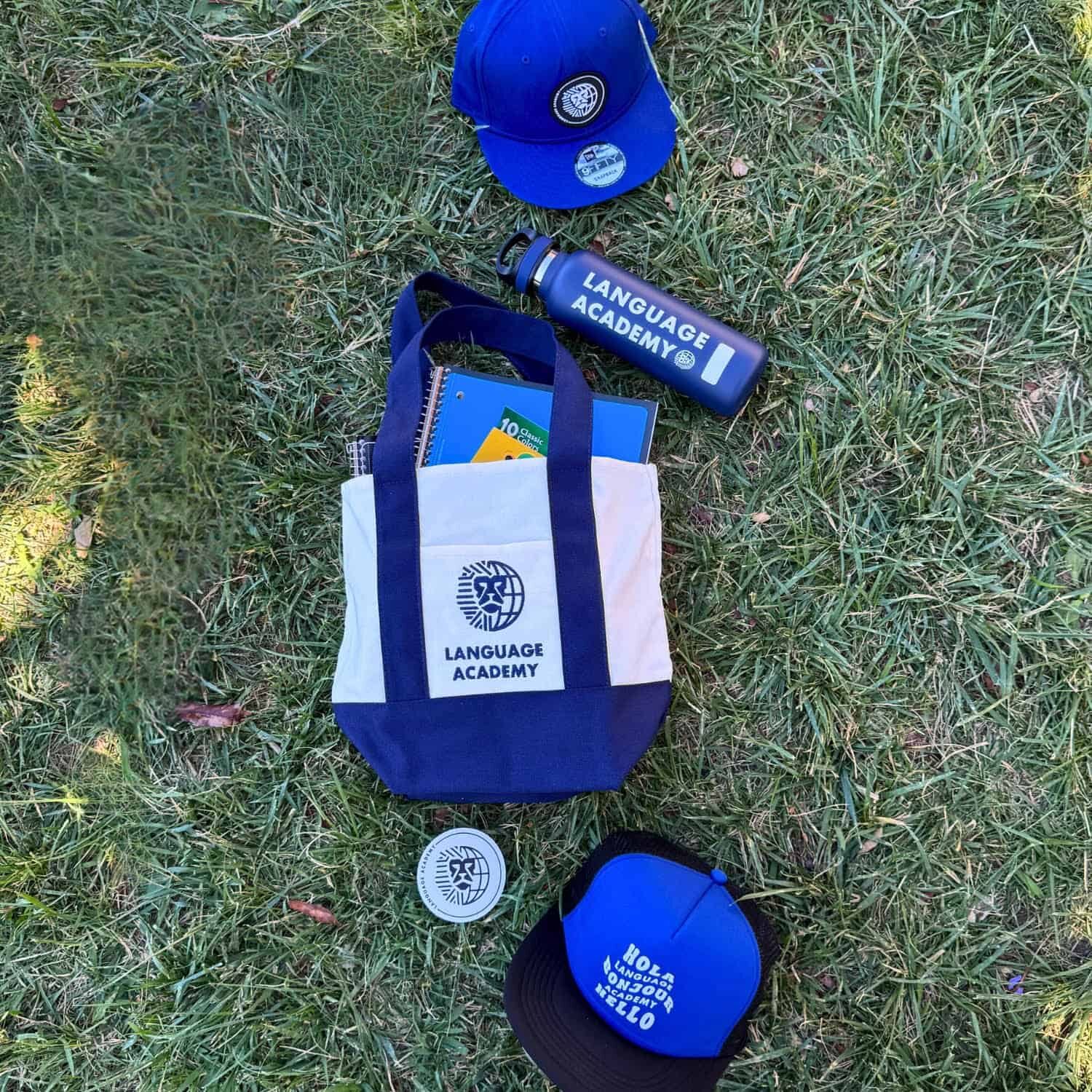 Language Academy merchandise, including tote bag, hat, water bottle, and sticker photographed against grass.